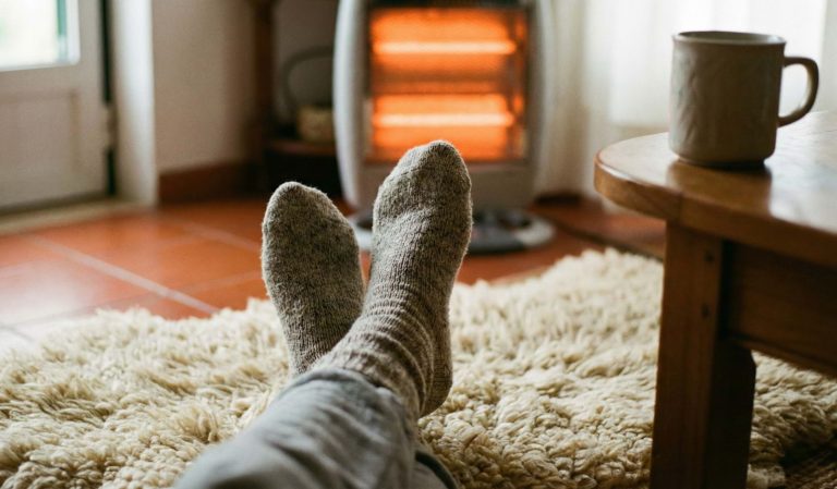 Pés com meias de lã grossas sobre um tapete felpudo em frente a um aquecedor elétrico, ilustrando como manter a casa quente e poupar energia no inverno.